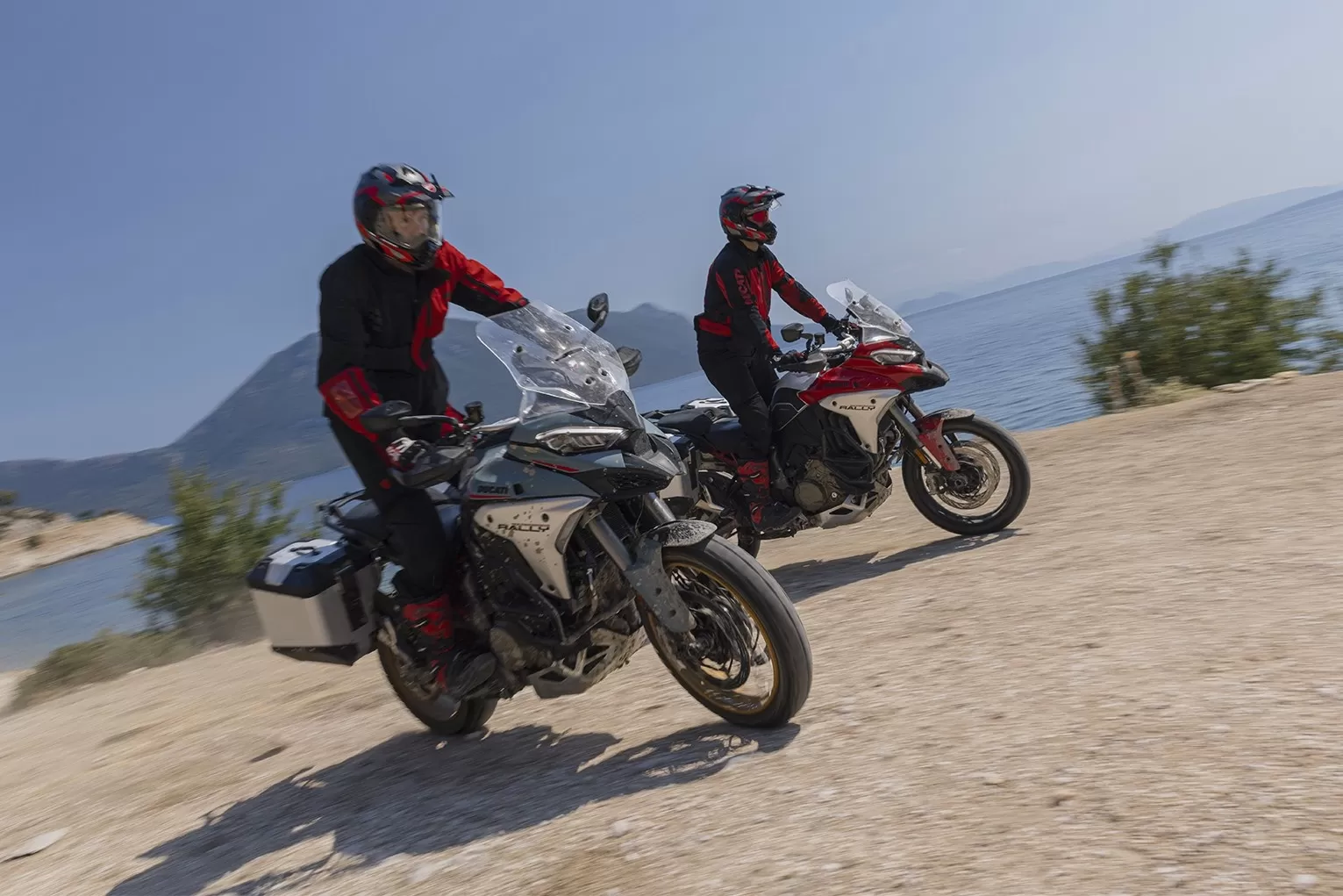 Two riders on new Ducati Multistrada V4 Rally motorcycles riding along a gravel trail in a scenic mountain landscape.